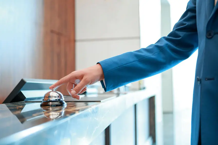 Hotel Add-On Kassensoftware Erweiterung: Person im blauen Jacket an einer Hotel-Rezeption läutet die Tischglocke am Tresen
