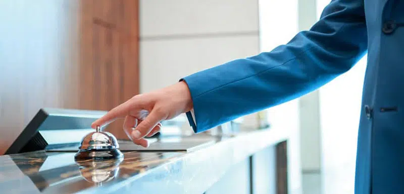 Person im Anzug in einem Hotel, vor dem Empfang, drückt auf eine Tischglocke bezugnehmend auf das GastroSoft Hotel Add-On